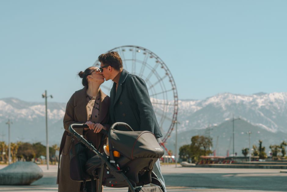 A couple kisses with a stroller in front of a Ferris wheel against a mountain backdrop.
