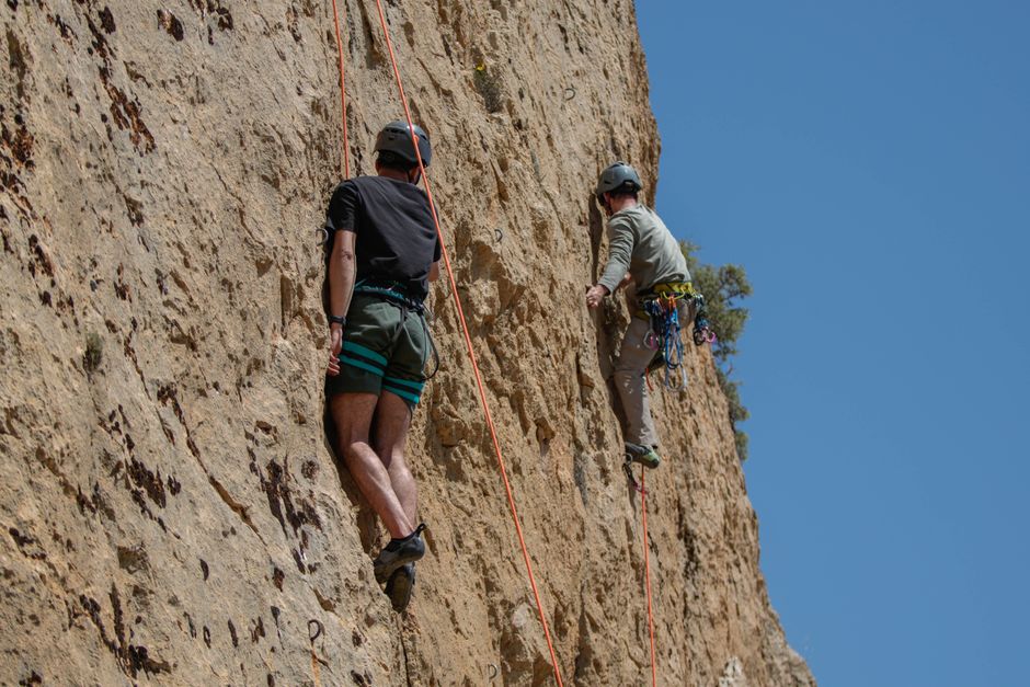 Two climbers ascend a rocky cliff with gear under a clear blue sky.