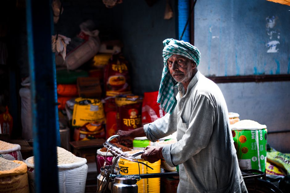 An elderly man stands with a bicycle in a vibrant Indian bazaar full of colorful goods.