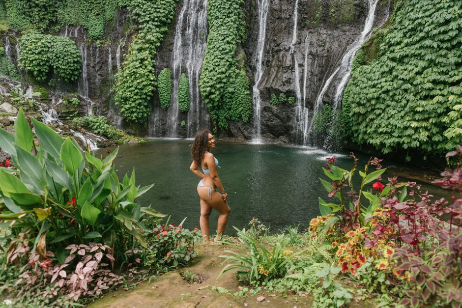 Side view of excited young ethnic female traveler in swimwear smiling while relaxing on lake shore near waterfall in tropical gorge