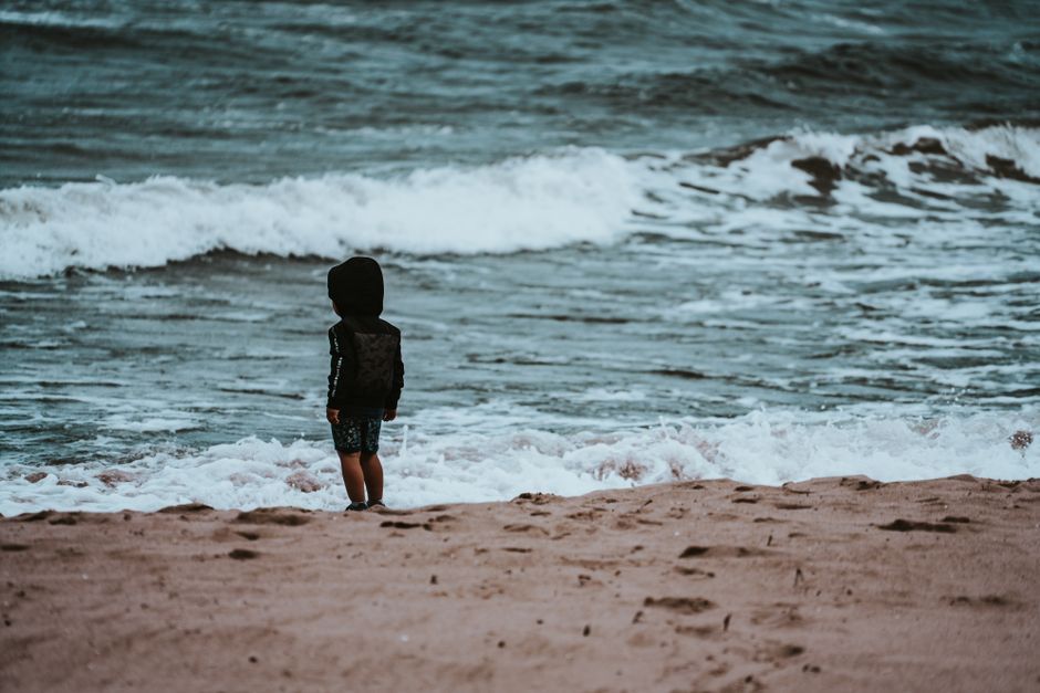 A child wearing a hoodie stands by the serene beach, gazing at the gentle waves.