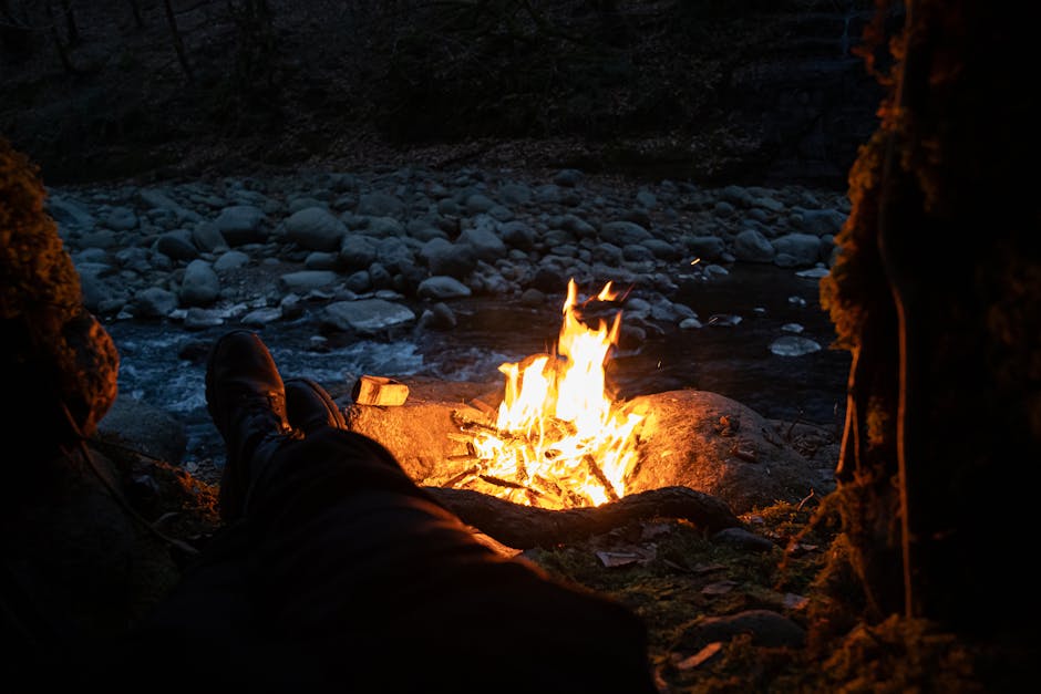 Relaxing by a campfire near a rocky river during twilight in a tranquil forest setting.