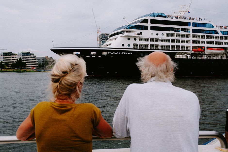 An elderly couple watches a large cruise ship in a bustling harbor.