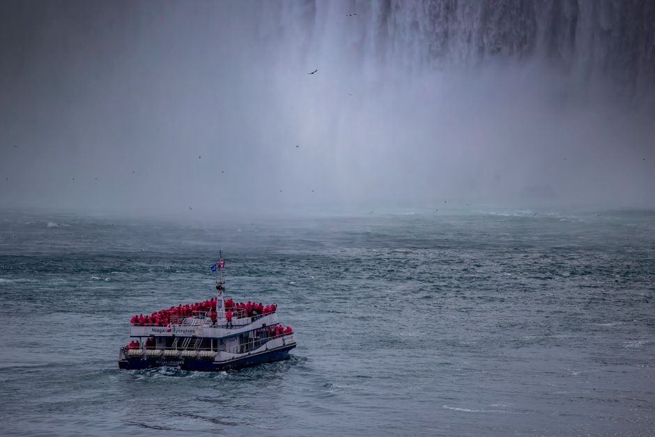 Tour boat approaches the majestic mist of Niagara Falls with passengers wearing red raincoats.