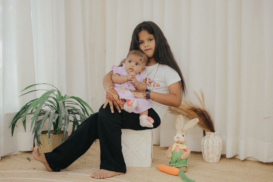 A young girl lovingly holds her baby sibling in a cozy indoor setting, surrounded by plants and decor.