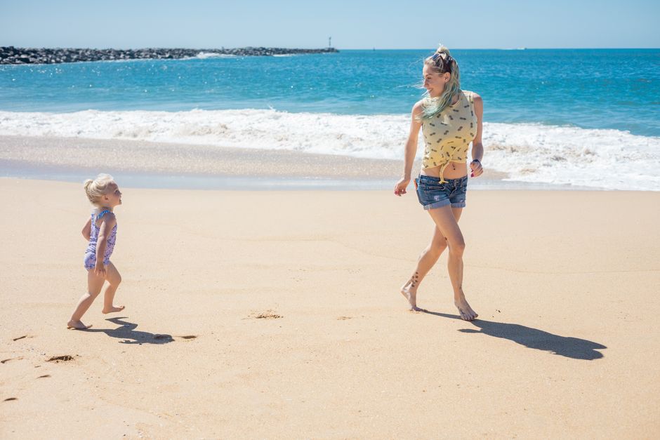 A mother and daughter running and playing on a sunny beach with waves in the background, enjoying family time.