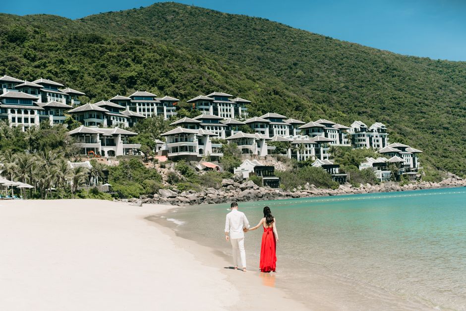 A couple walks hand in hand along a scenic beach next to luxurious villas, enjoying a sunny day.