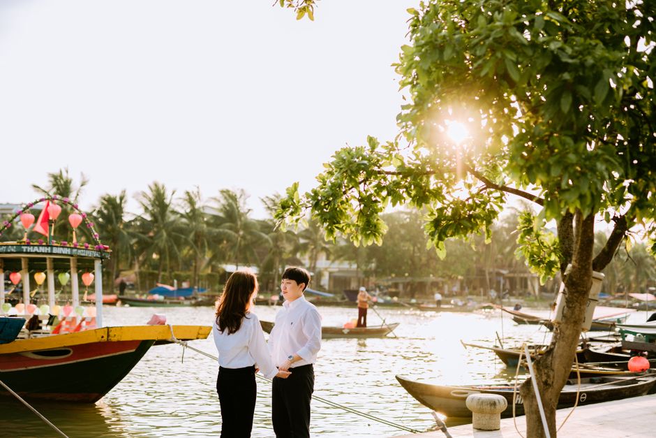 A couple shares a tender moment by a sunny riverside harbor with boats and palm trees in the background.
