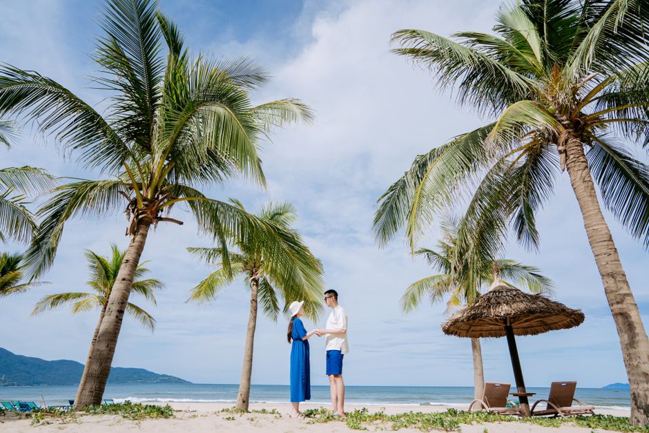 A couple holds hands on a sunny beach surrounded by coconut trees, enjoying a tropical vacation.