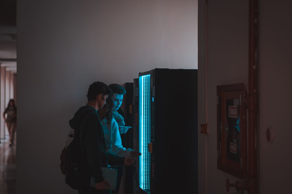 Group of teenagers using a vending machine in a school corridor, captured in ambient lighting.