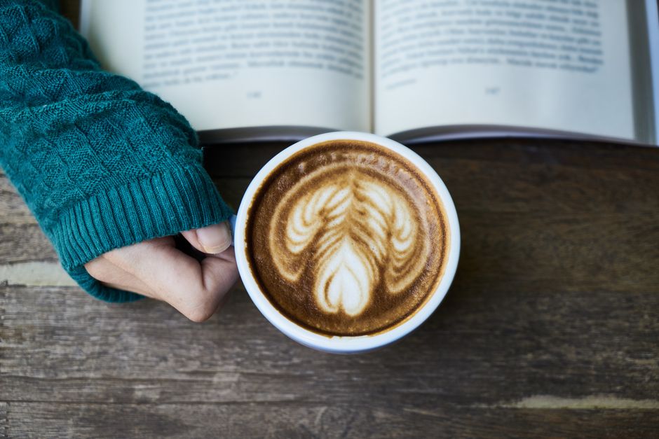 A warm latte with artistic foam held next to an open book on a wooden table, perfect for a cozy moment.