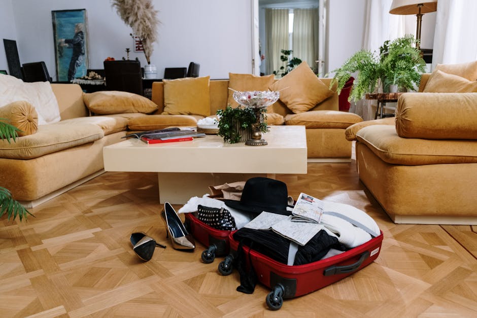 A stylish living room with beige couches and an open suitcase on a parquet floor, surrounded by plants.