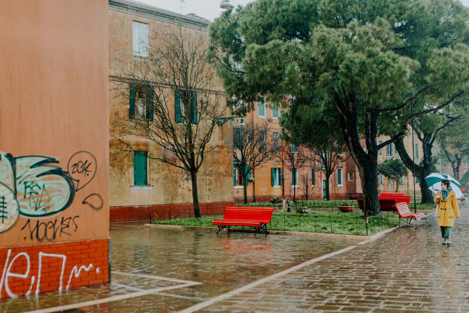An urban street scene with graffiti, benches, and a person walking in the rain under a blue umbrella.