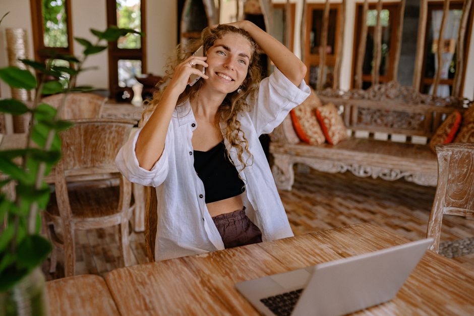 Woman enjoying a phone conversation while sitting at a wooden table with a laptop.