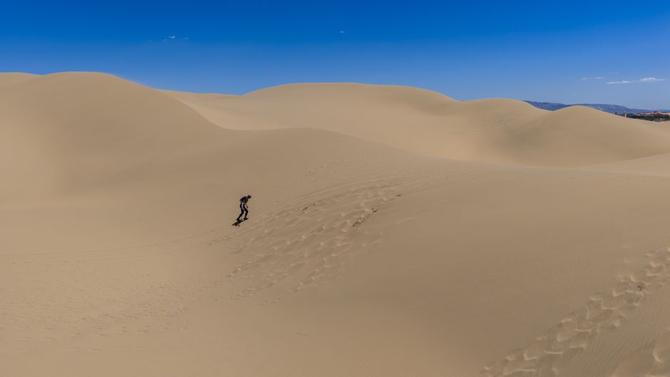 A lone sandboarder descends vast desert dunes under clear blue skies.