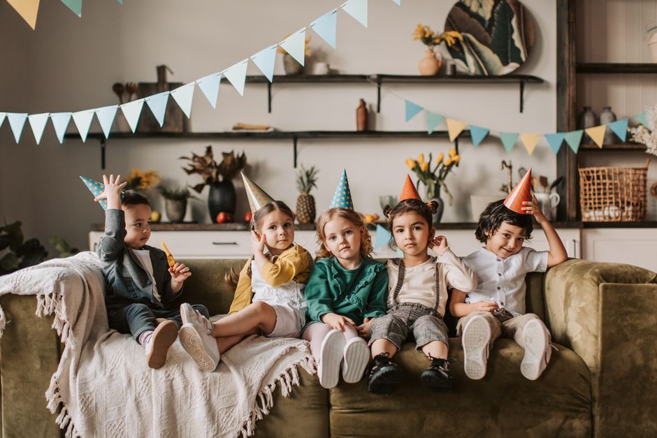 Children enjoying a birthday party indoors, wearing colorful hats and sitting on a sofa with decorations.